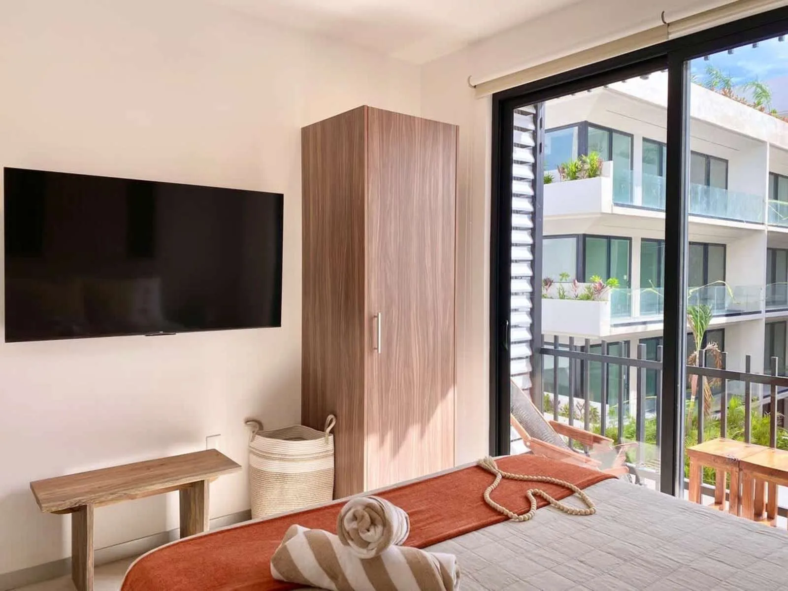 Un dormitorio luminoso tiene un televisor montado en la pared y un balcón con vistas a edificios vecinos.