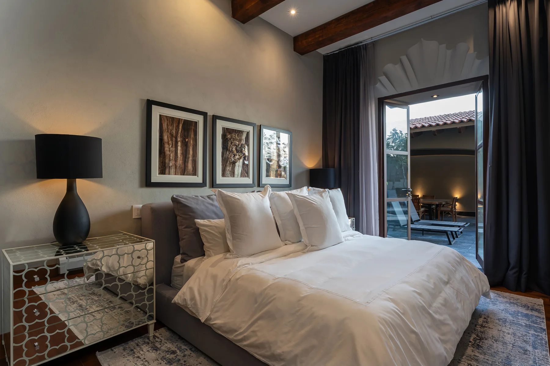 A bedroom with a neatly made bed, framed artwork, and dark curtains around a glass door.