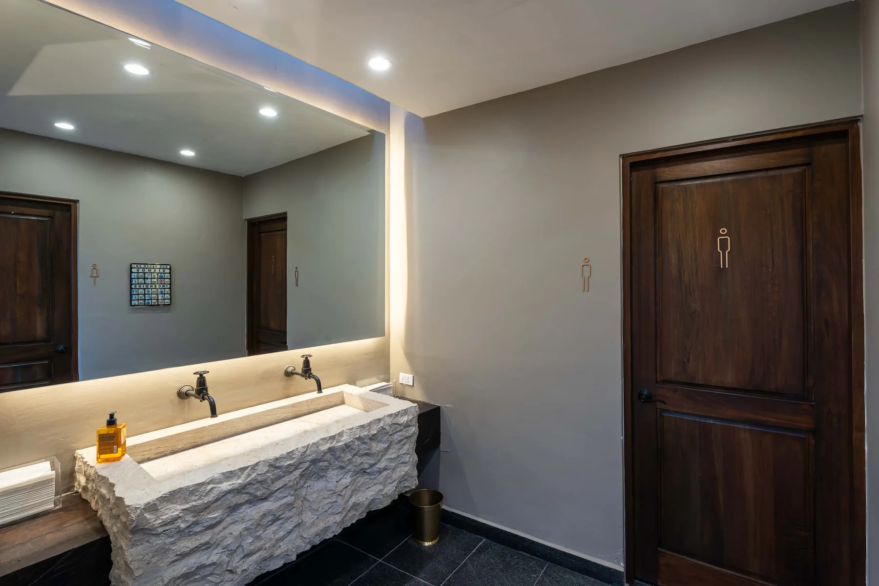 A modern bathroom with a stone vanity, wall mirror, and dark wooden doors.