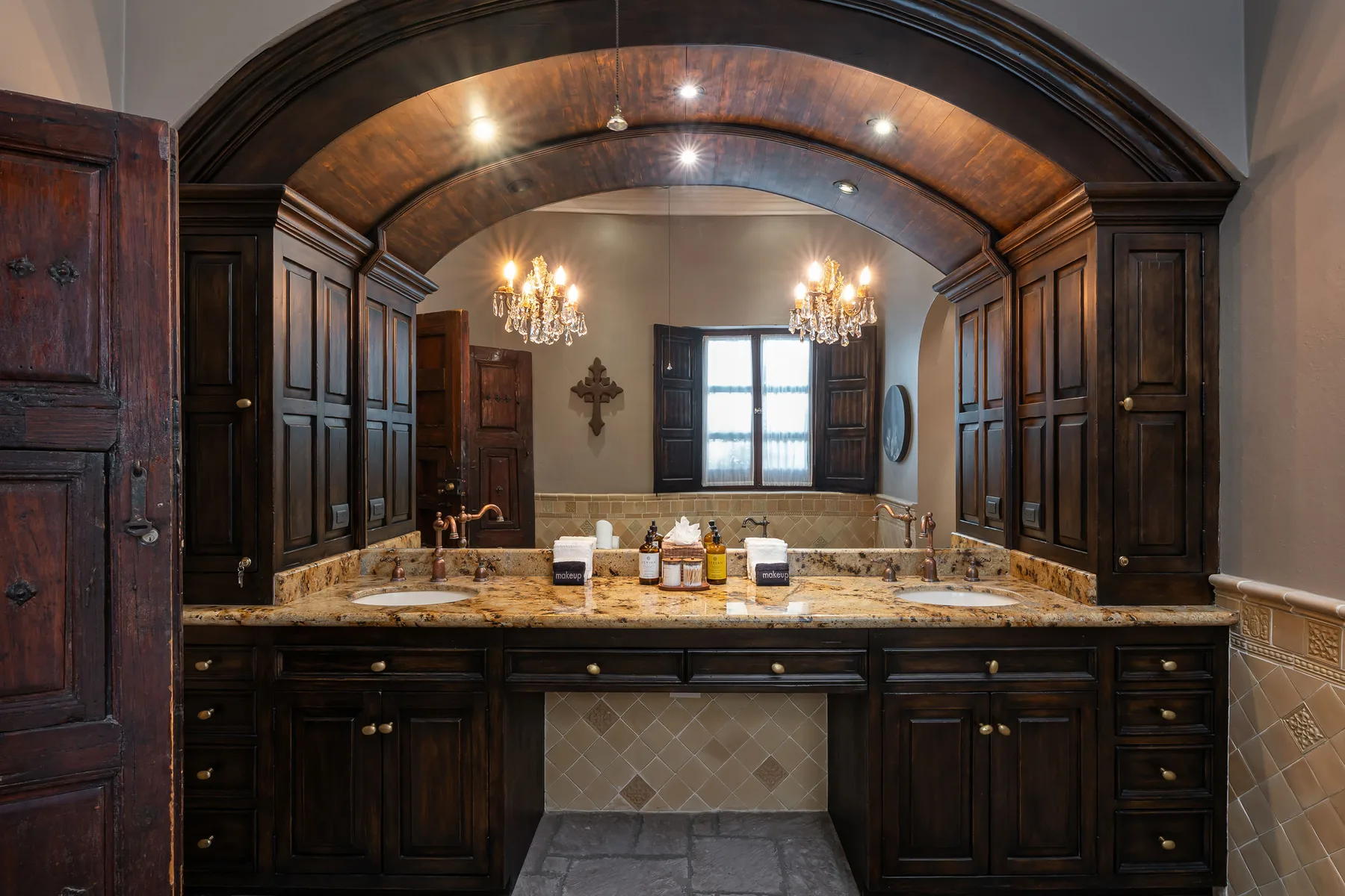 A dark wood bathroom vanity sits beneath an arched ceiling, with a window beyond.