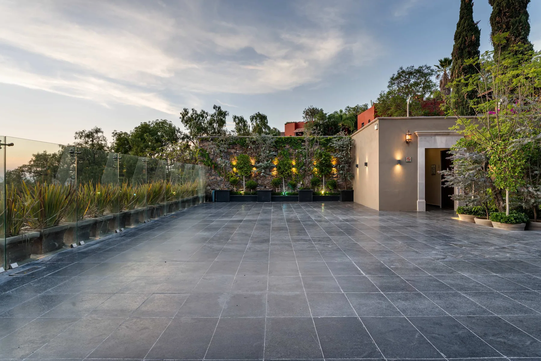 A spacious tiled outdoor terrace bordered by glass railings and greenery.