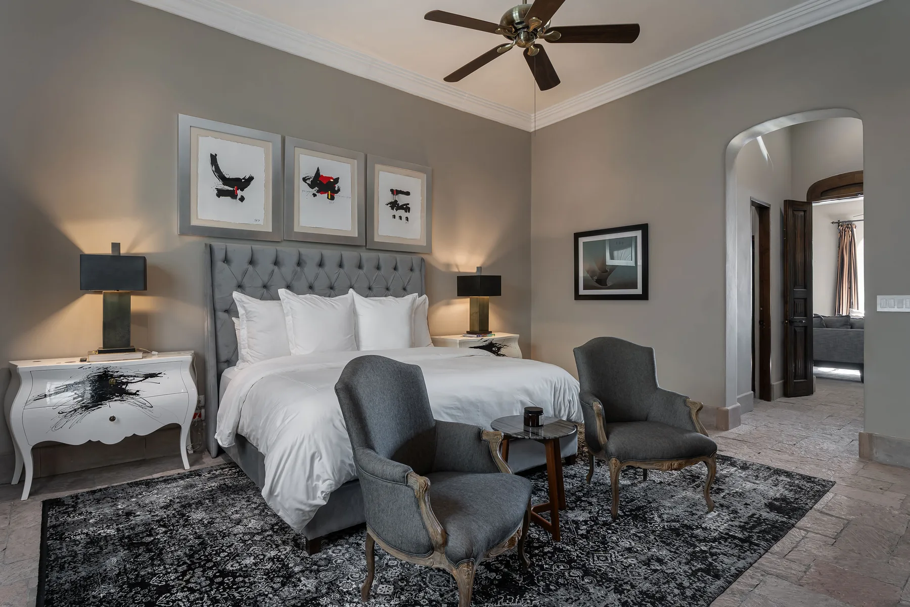 A bedroom with a bed, two chairs, and framed artwork above the headboard.