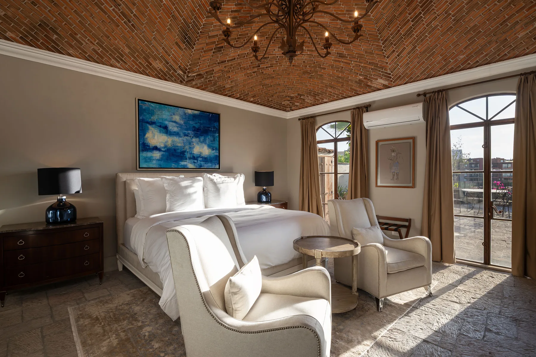 A bright bedroom with a large bed, seating, and tall windows beneath a brick ceiling.