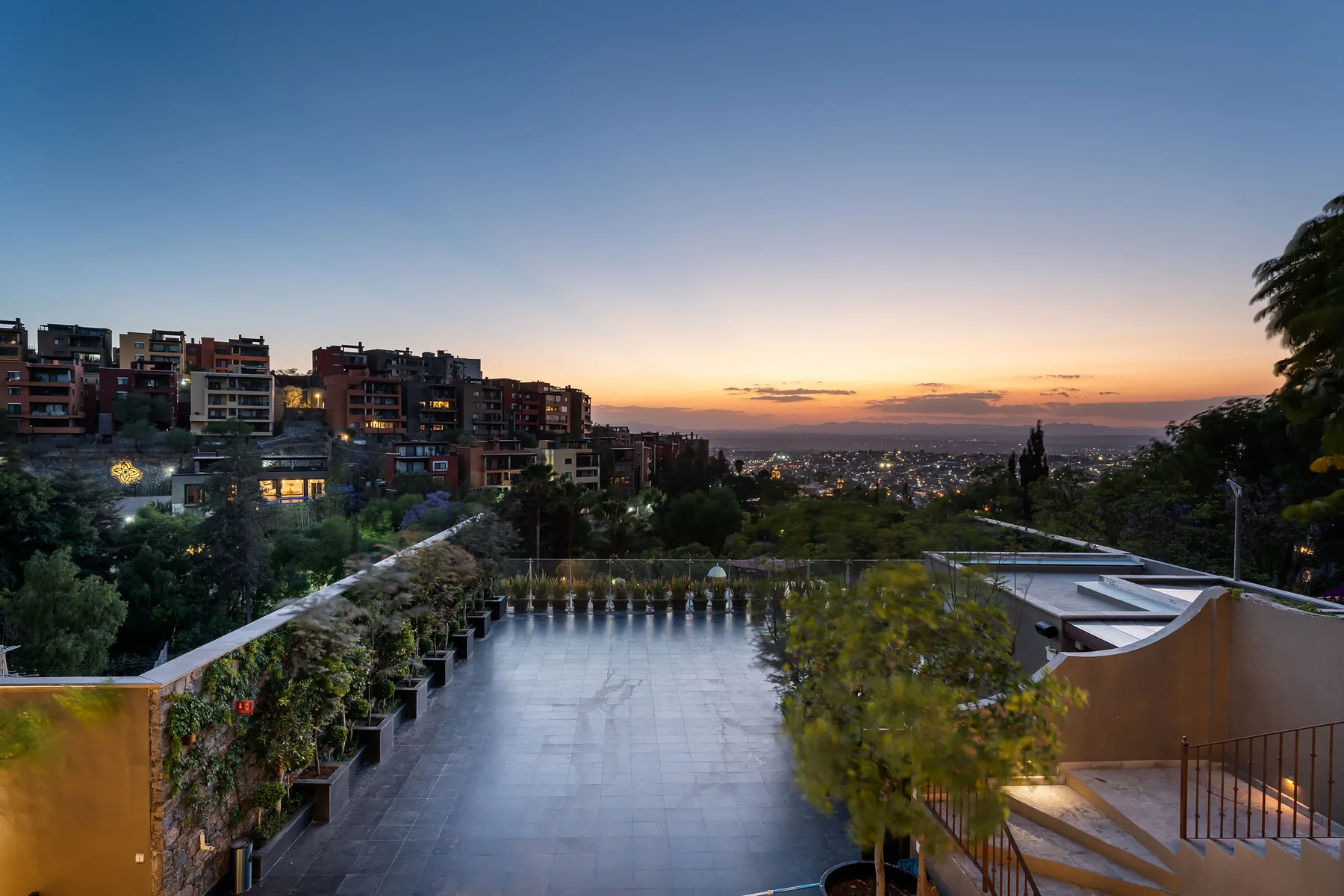 A rooftop terrace overlooks hillside buildings and a city at sunset.