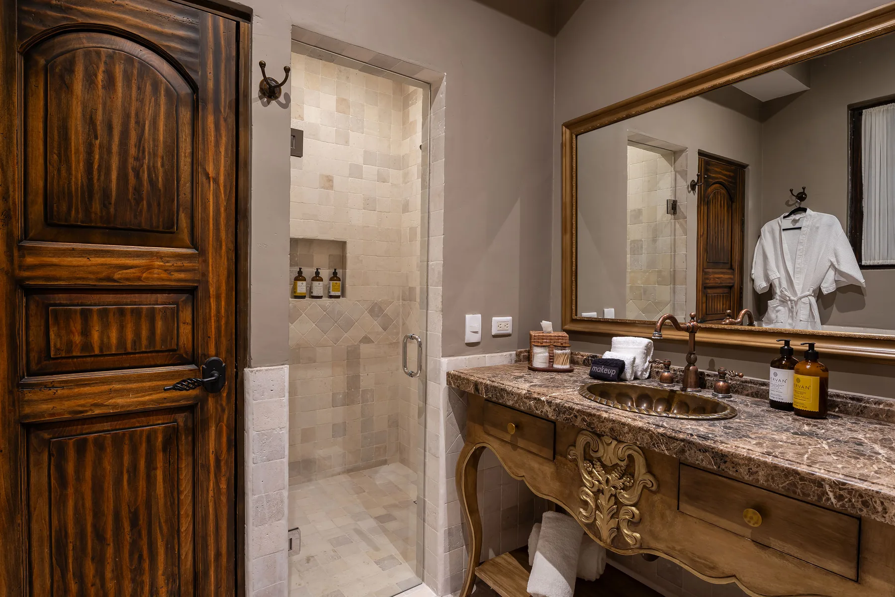 A bathroom vanity and mirror sit beside a tiled shower and dark wooden door.