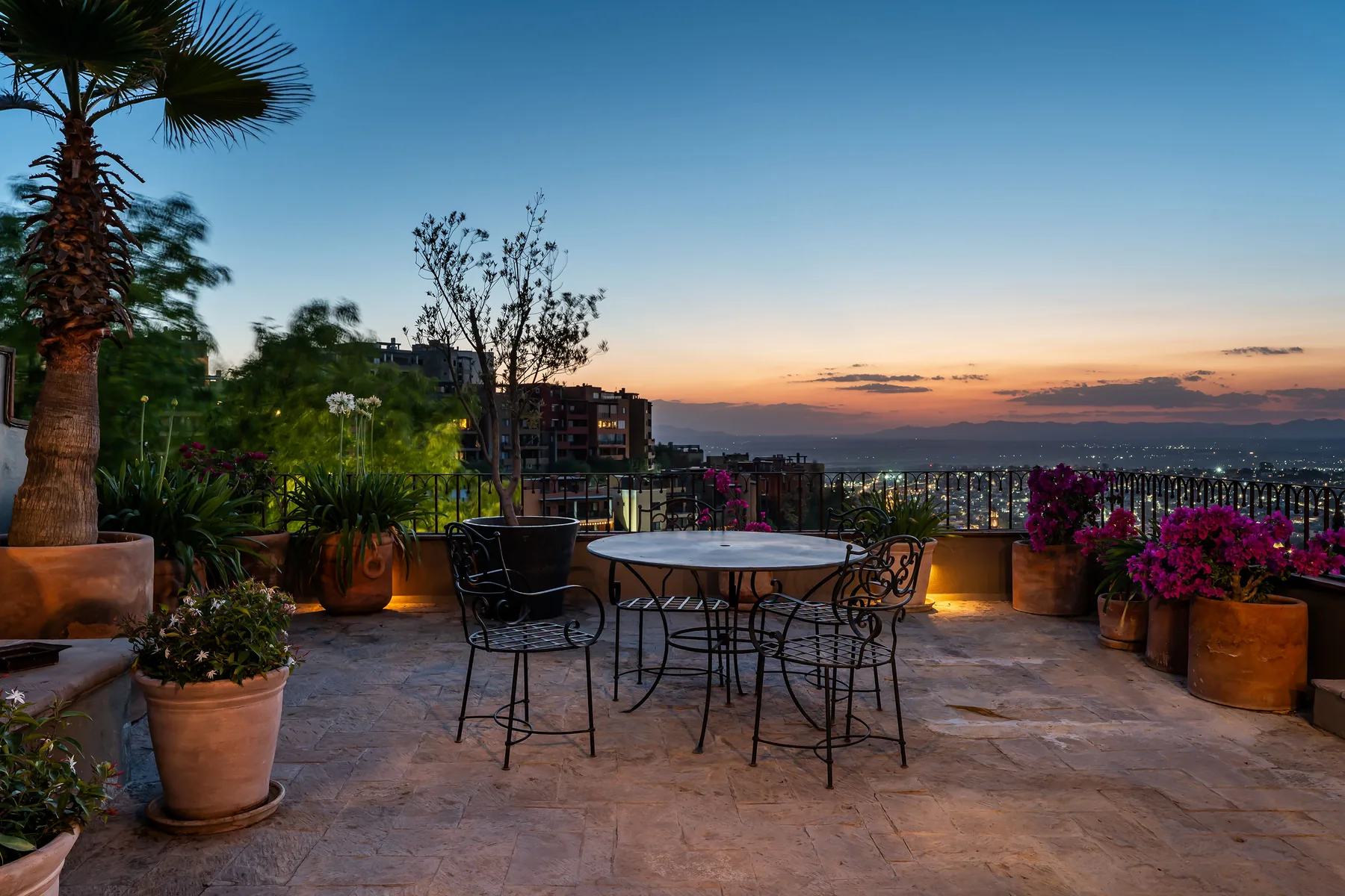 An outdoor terrace with a round table, seating, and potted plants overlooks the sunset.