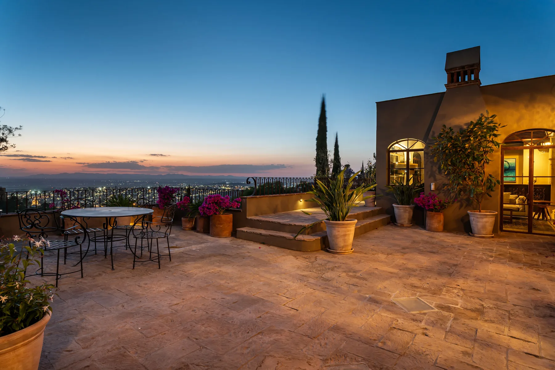 A rooftop terrace with outdoor seating, potted plants, and a city view at dusk.