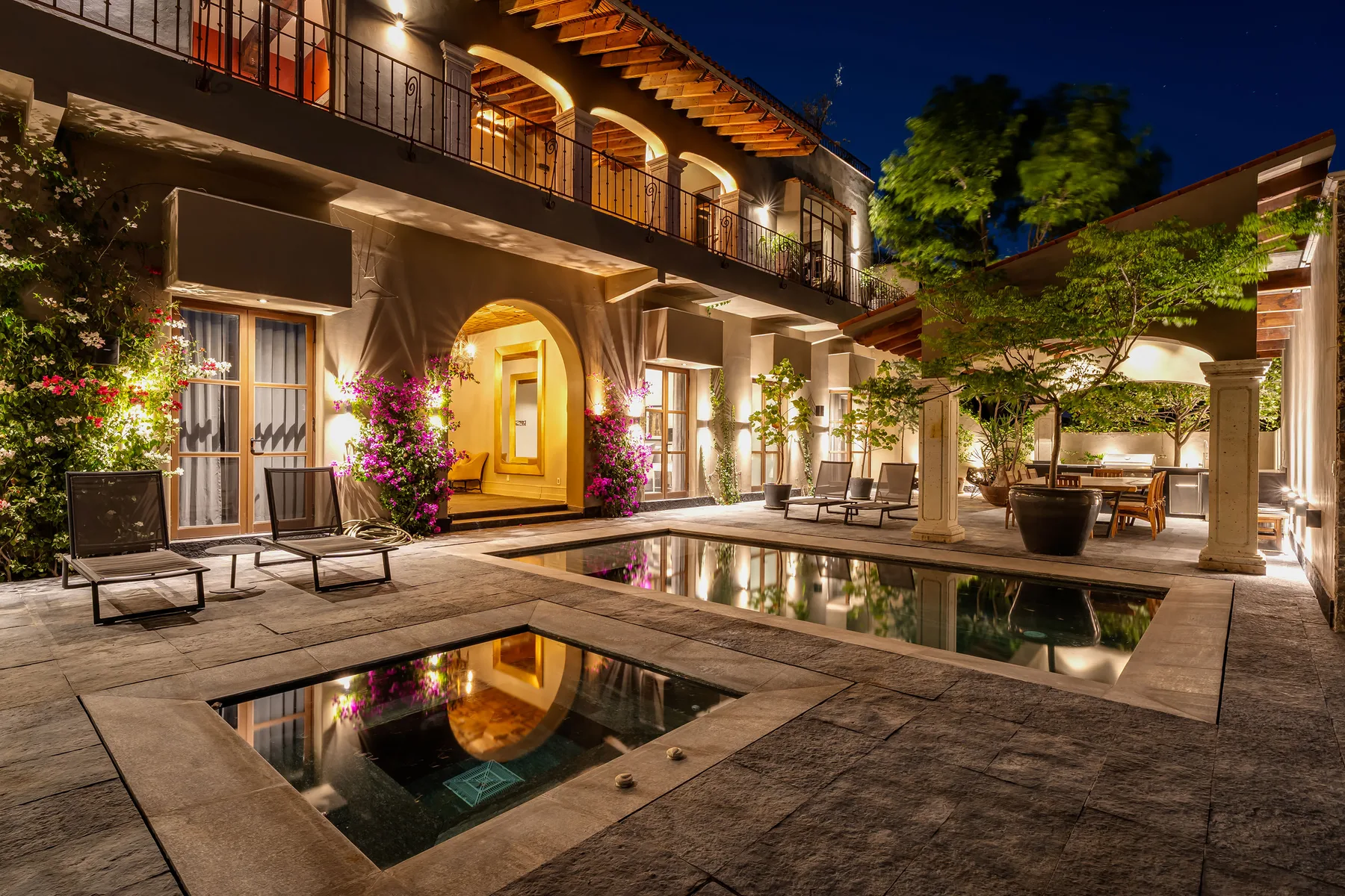 A lit courtyard patio with seating surrounds a swimming pool beside a two-story building.