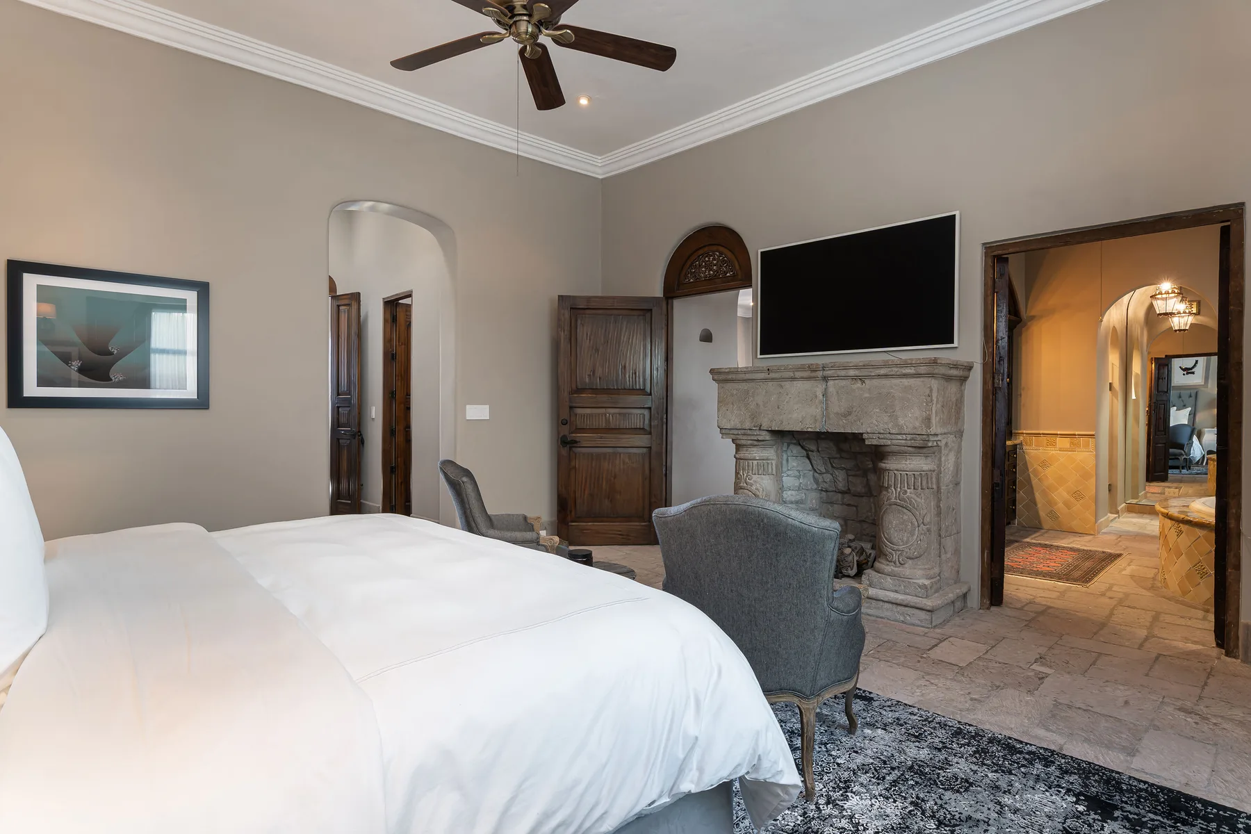 A bedroom with a large bed, seating, and a stone fireplace beneath a television.
