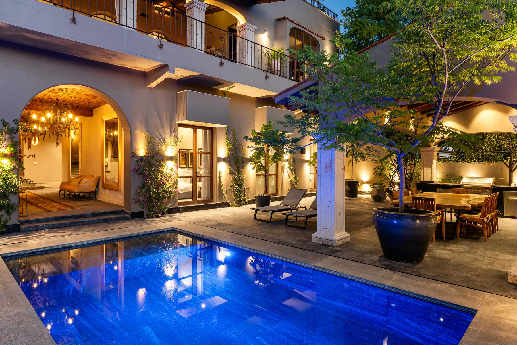 A courtyard pool glows beside a dining area and an arched entrance at dusk.