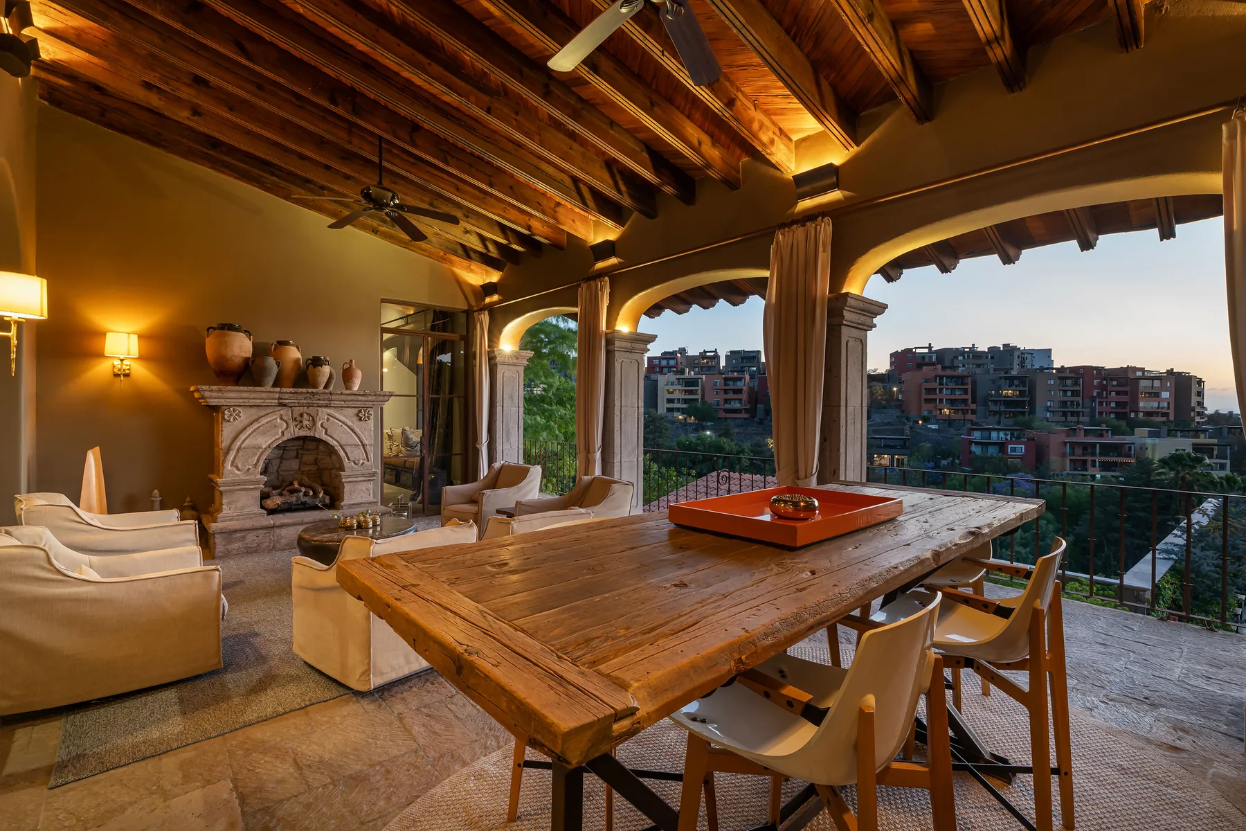 A covered terrace features a large wooden dining table and seating overlooking nearby buildings.