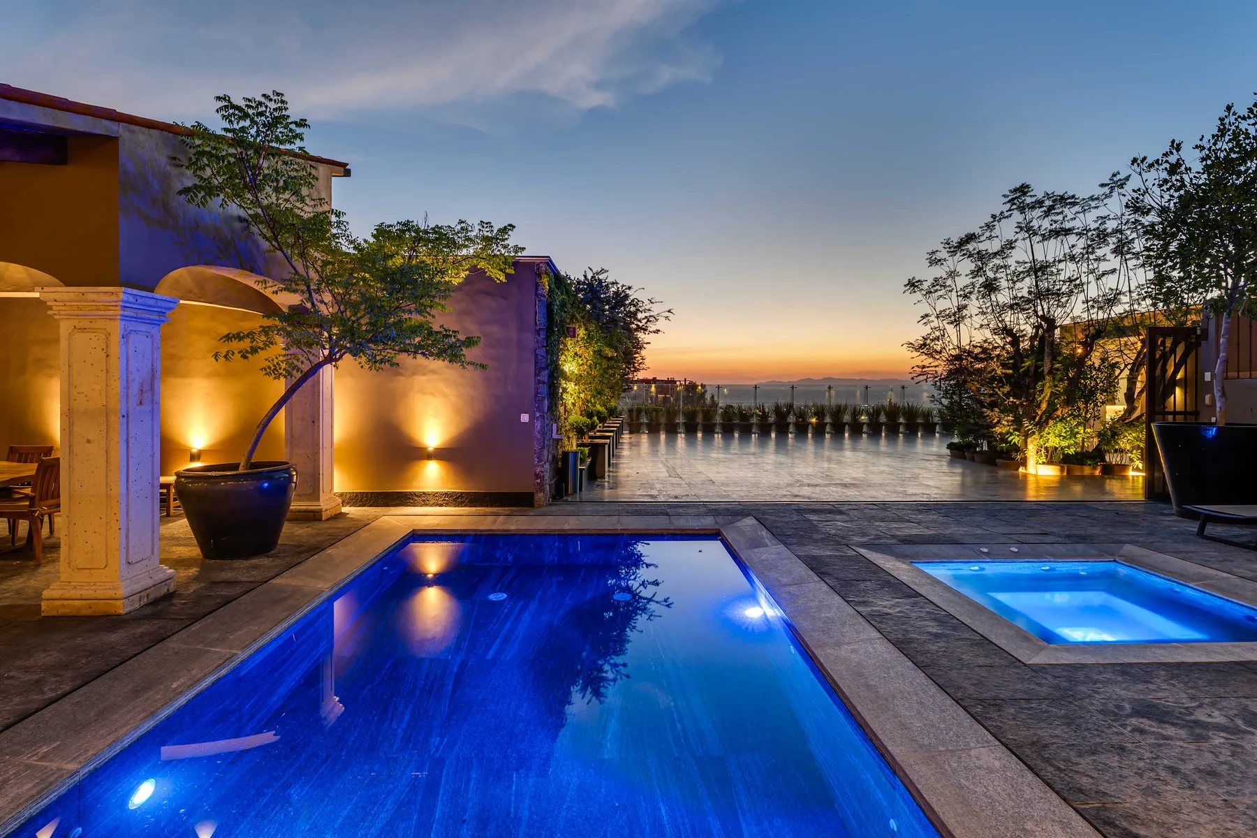 An outdoor swimming pool and smaller spa sit beside a terrace at sunset.