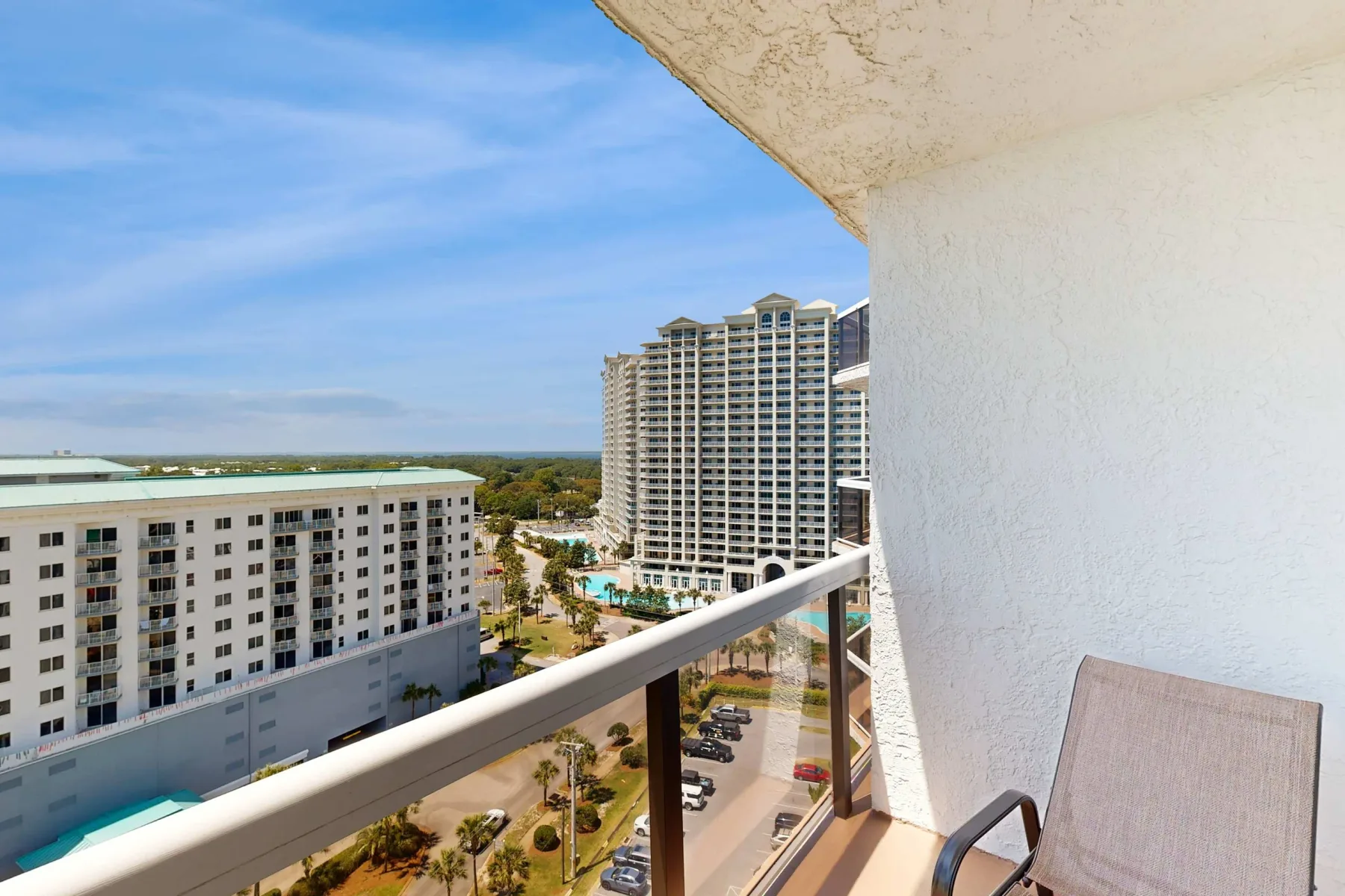 A balcony overlooks nearby buildings, palm trees, and a pool.