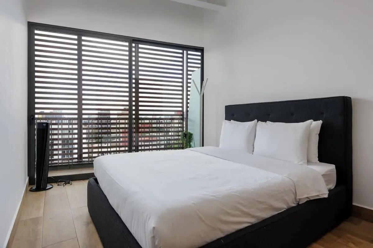 A bedroom with a white bed and large slatted window blinds.