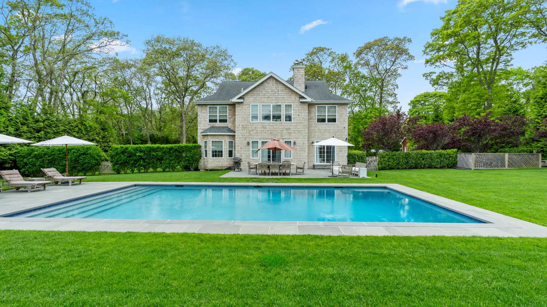 An outdoor swimming pool sits in front of a two-story house and green lawn.