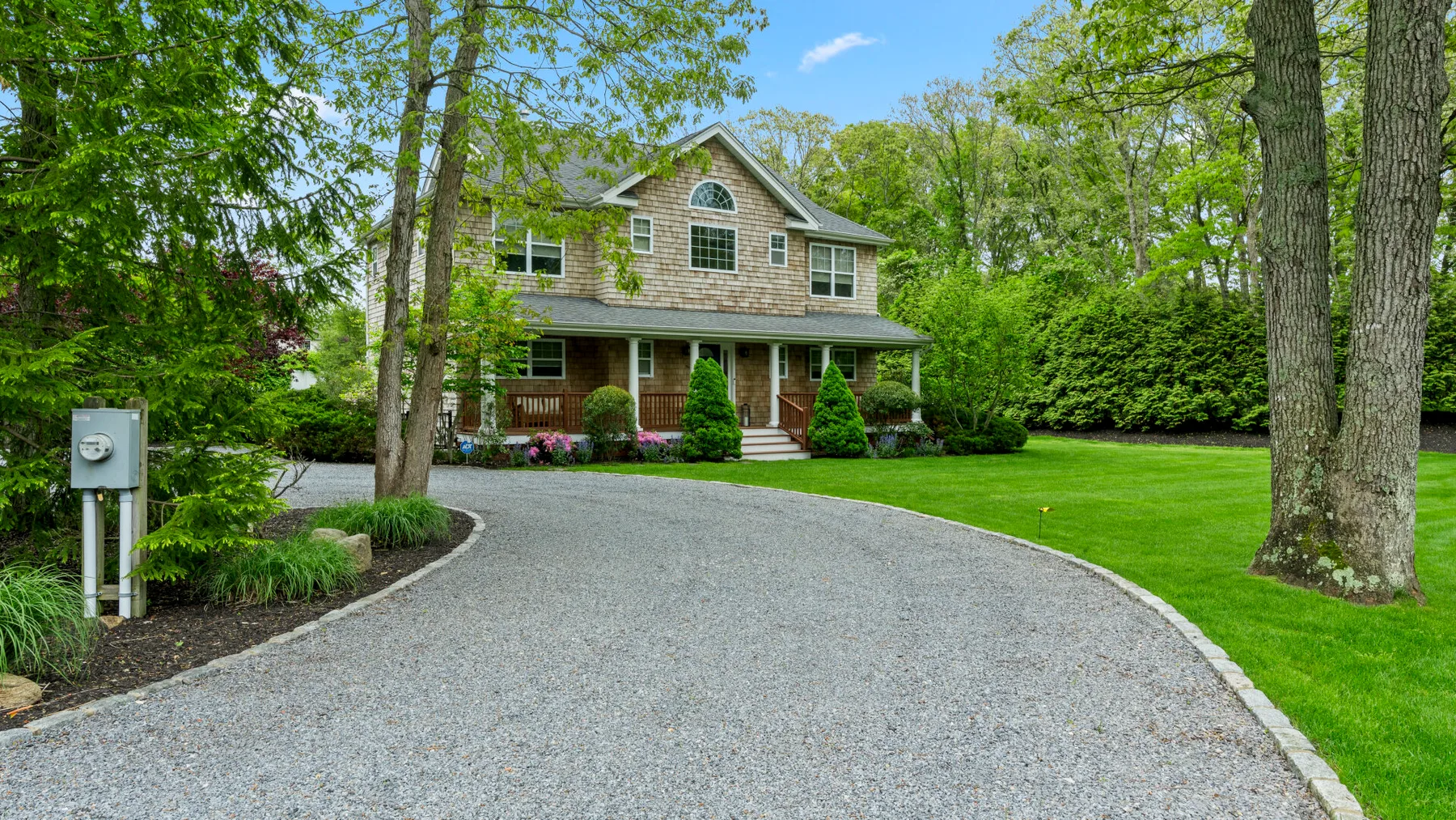 A shingle-sided house stands beyond a curved gravel driveway, surrounded by trees and lawn.