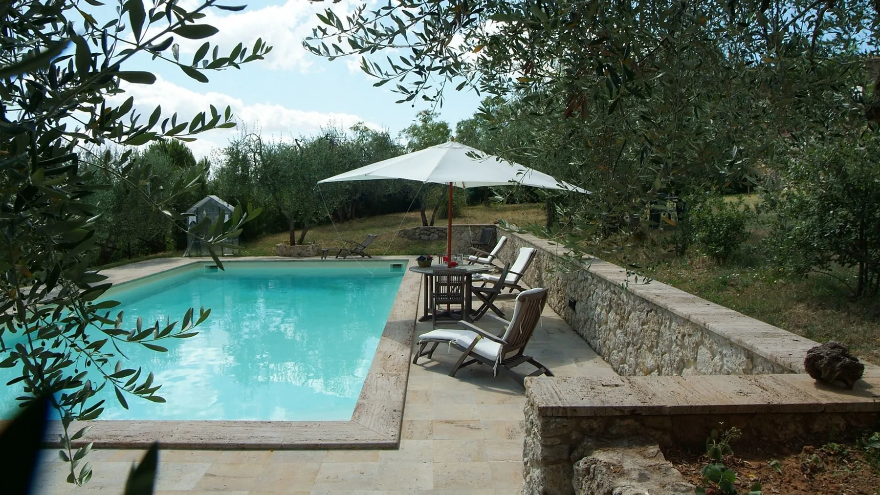 Una piscina al aire libre junto a un patio de piedra con asientos y una sombrilla.