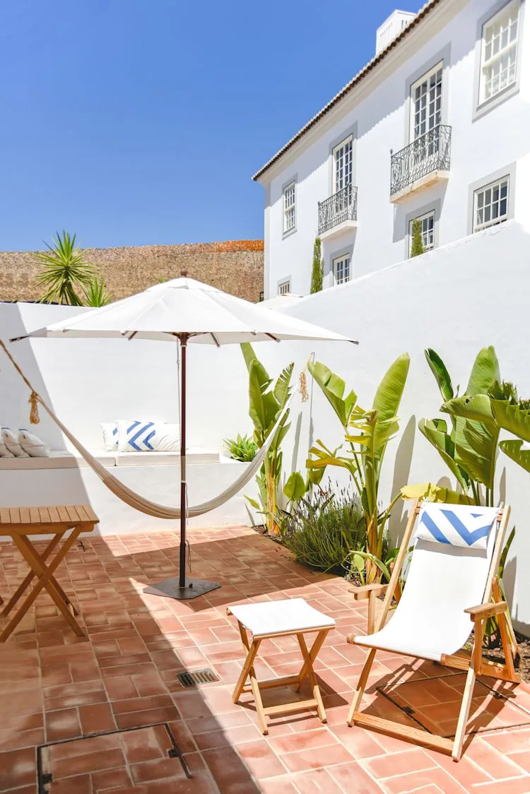 Espacio representativo del hotel: Un patio soleado con suelo de baldosas, tumbonas, una sombrilla y plantas tropicales.
