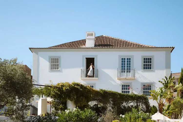 Espacio representativo del hotel: Un edificio blanco con tejado de tejas se alza detrás de plantas frondosas.