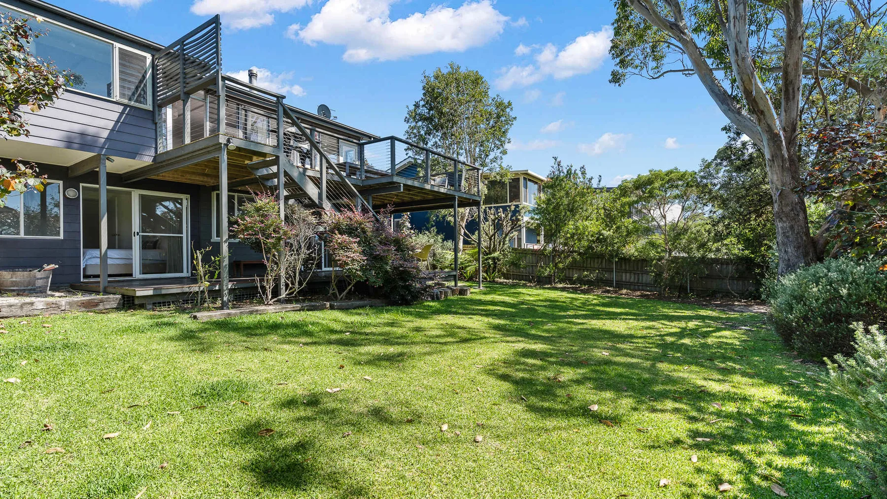 Un patio trasero cubierto de césped junto a una casa moderna, con una terraza elevada y árboles frondosos.