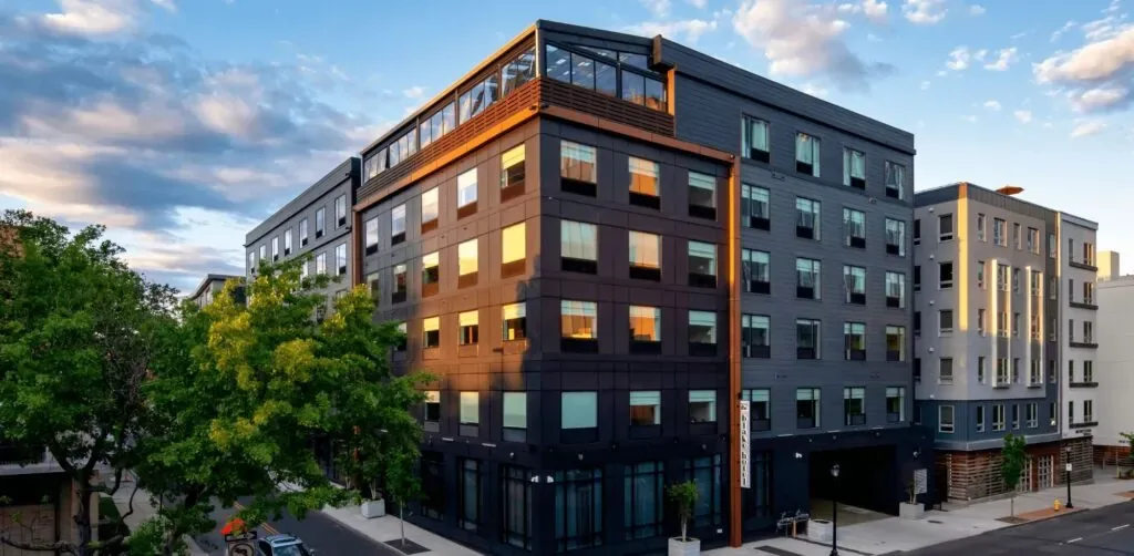 Representative hotel space: Modern multi-story building with dark and brick-colored exterior sections beside a tree-lined street.