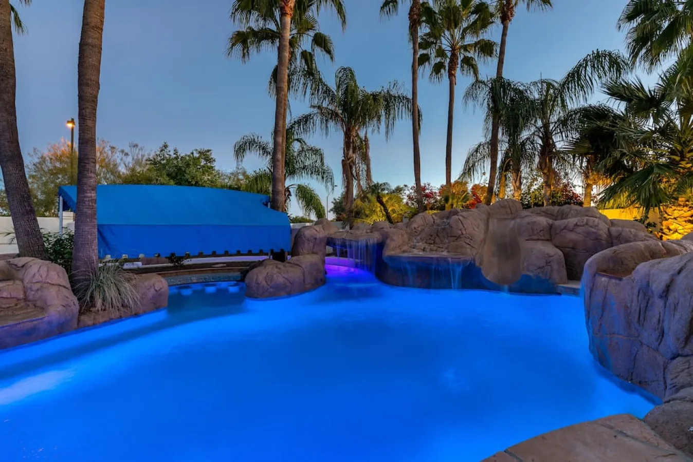 Una piscina iluminada de azul está rodeada de palmeras y formaciones rocosas.