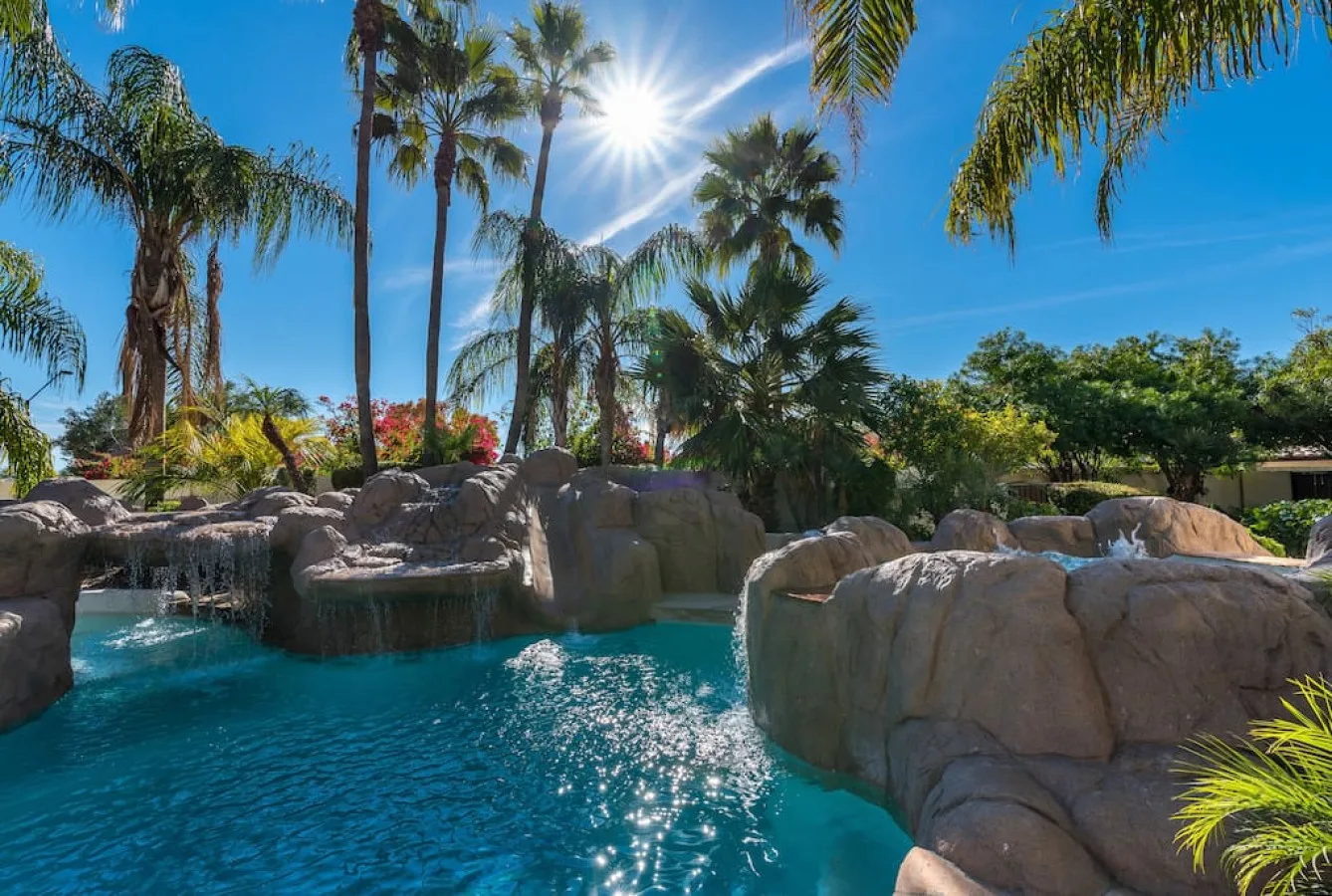 Una piscina al aire libre, iluminada por el sol, está rodeada de palmeras y grandes rocas.