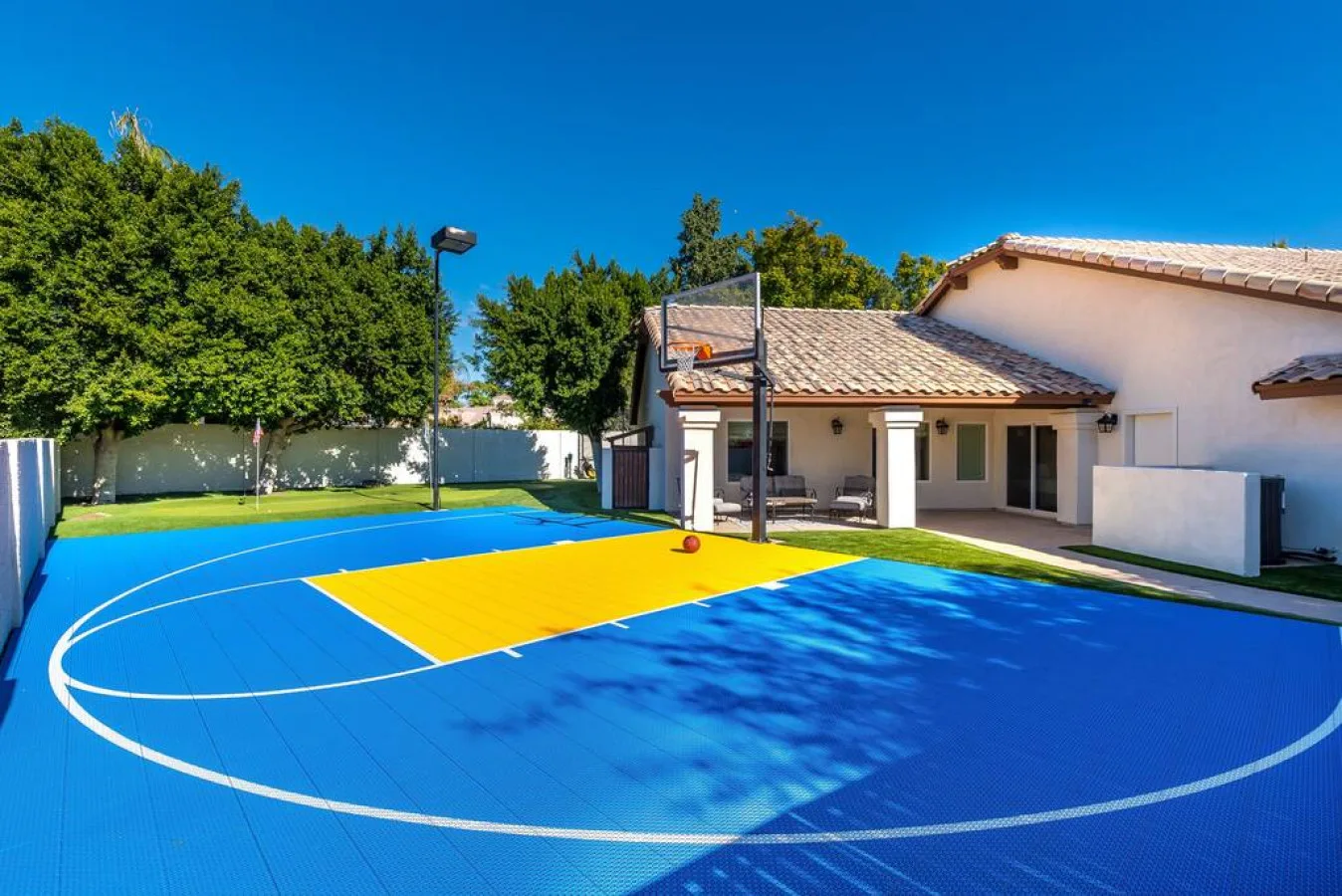 Cancha de baloncesto al aire libre junto a una casa, con superficies de juego azules y amarillas.