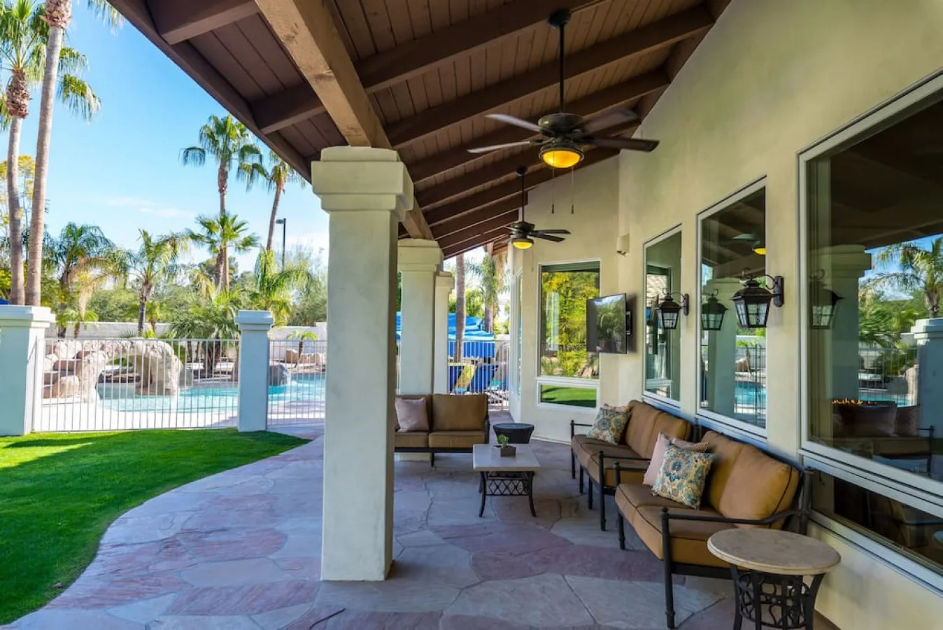 Un patio cubierto tiene asientos junto a grandes ventanas, con una piscina visible más allá del césped.