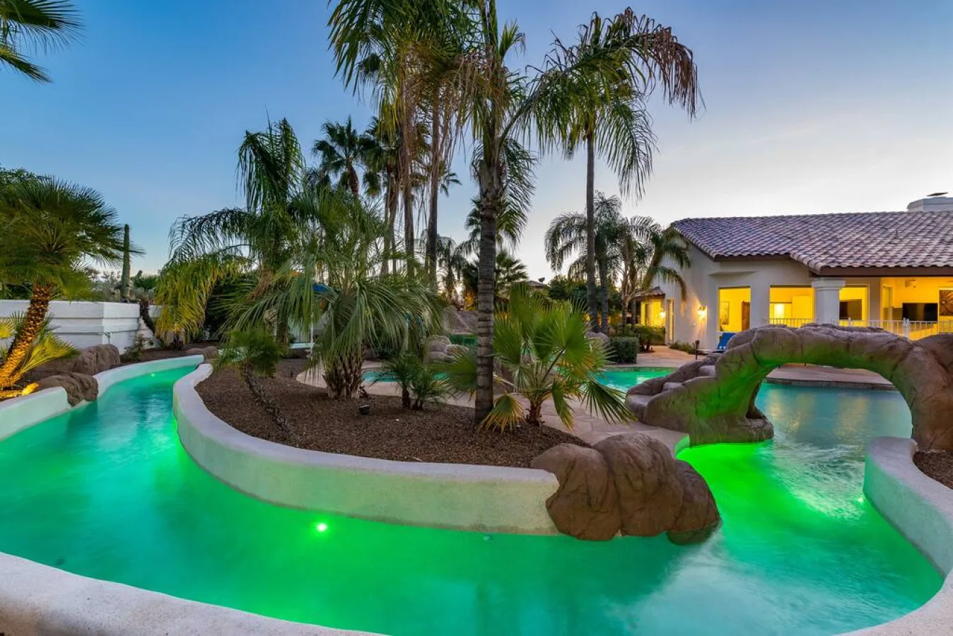 Una piscina ajardinada serpentea entre palmeras junto a un edificio iluminado.