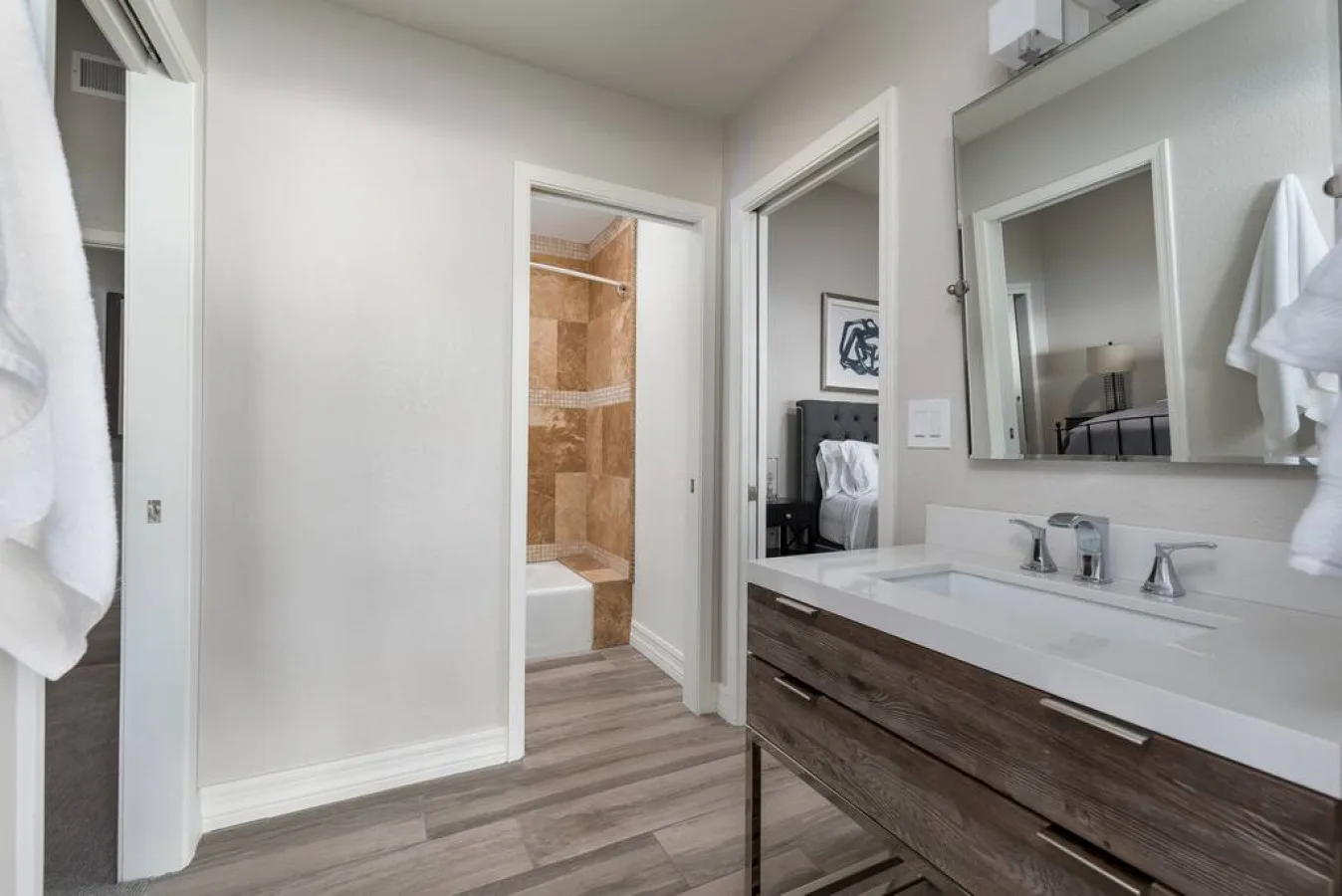 Un tocador de baño con un espejo amplio está junto a una puerta abierta que da a una ducha con azulejos.