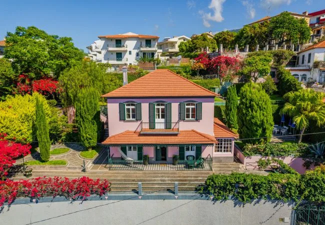 A pink two-story house with a tiled roof stands among landscaped greenery.