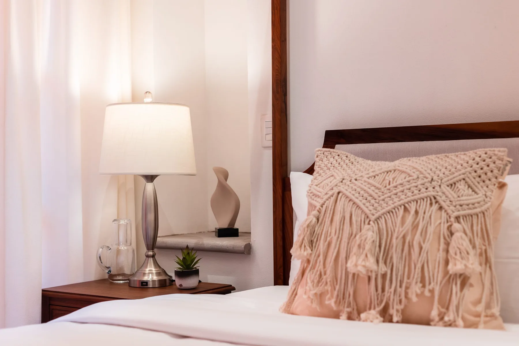 A bedroom with a bed, fringed decorative pillow, and bedside lamp.