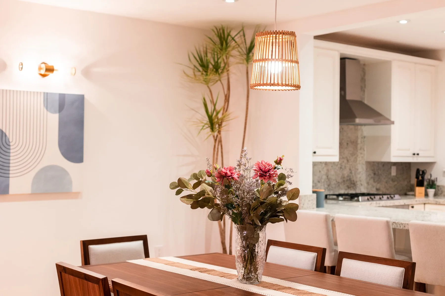 A dining area with a wooden table, flowers, and an open kitchen.