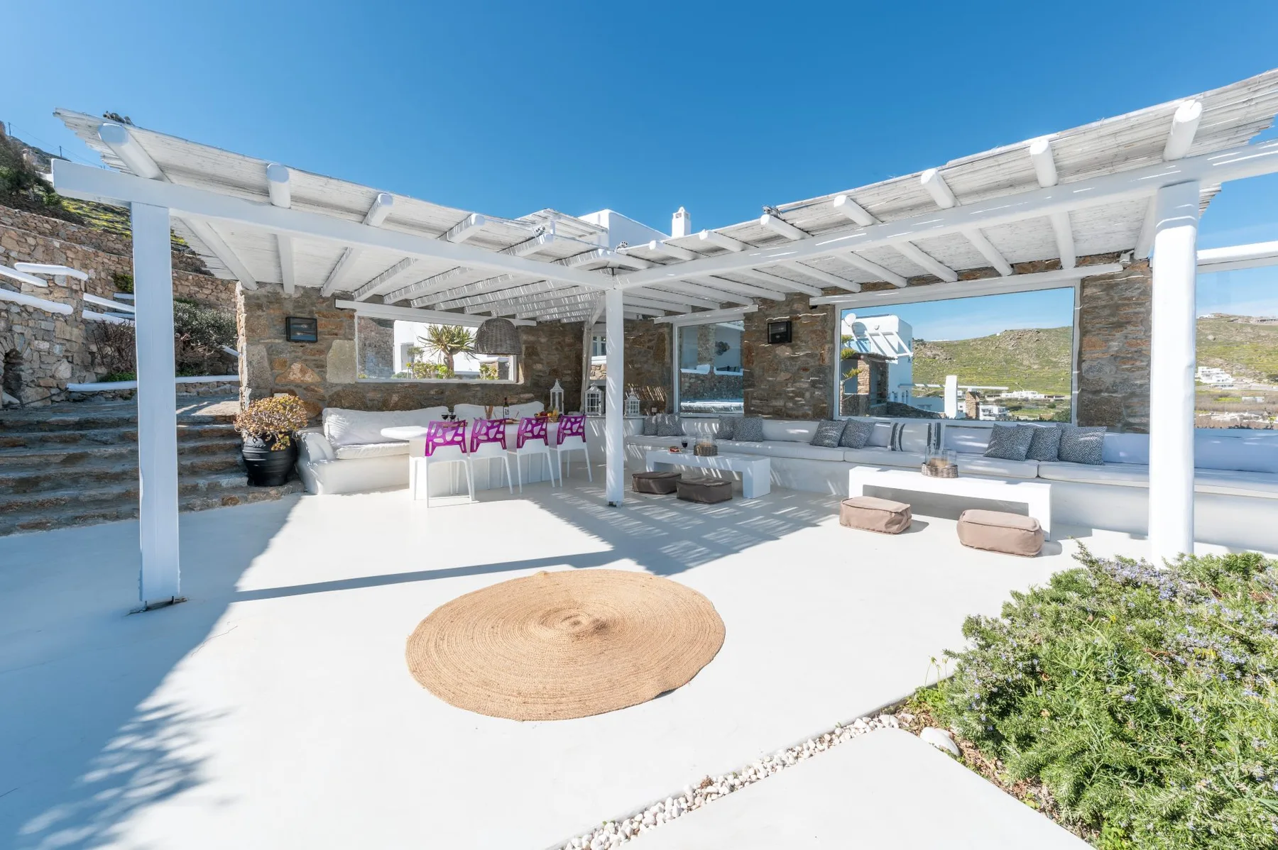 Un patio al aire libre con asientos blancos y una pérgola da a una ladera verde.