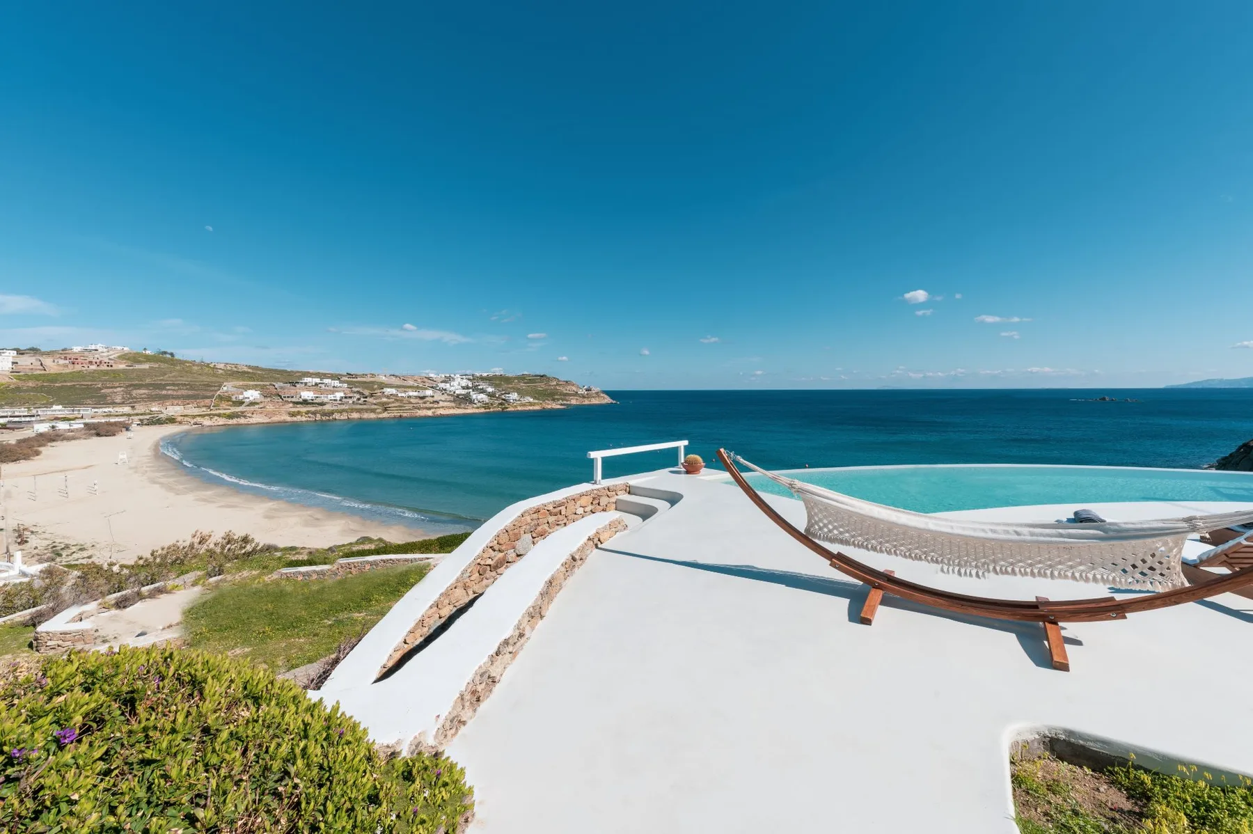 Una terraza en la azotea domina una playa curva y el océano azul, con una hamaca en primer plano.