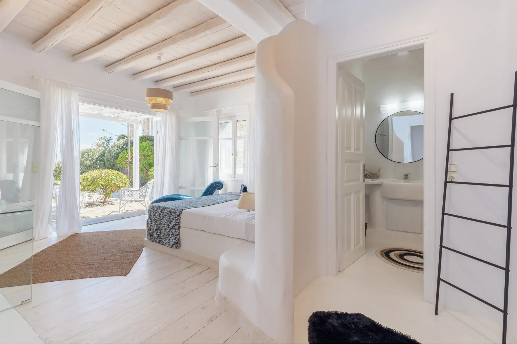 Un dormitorio luminoso se abre a un baño, con una cama y un espejo redondo.