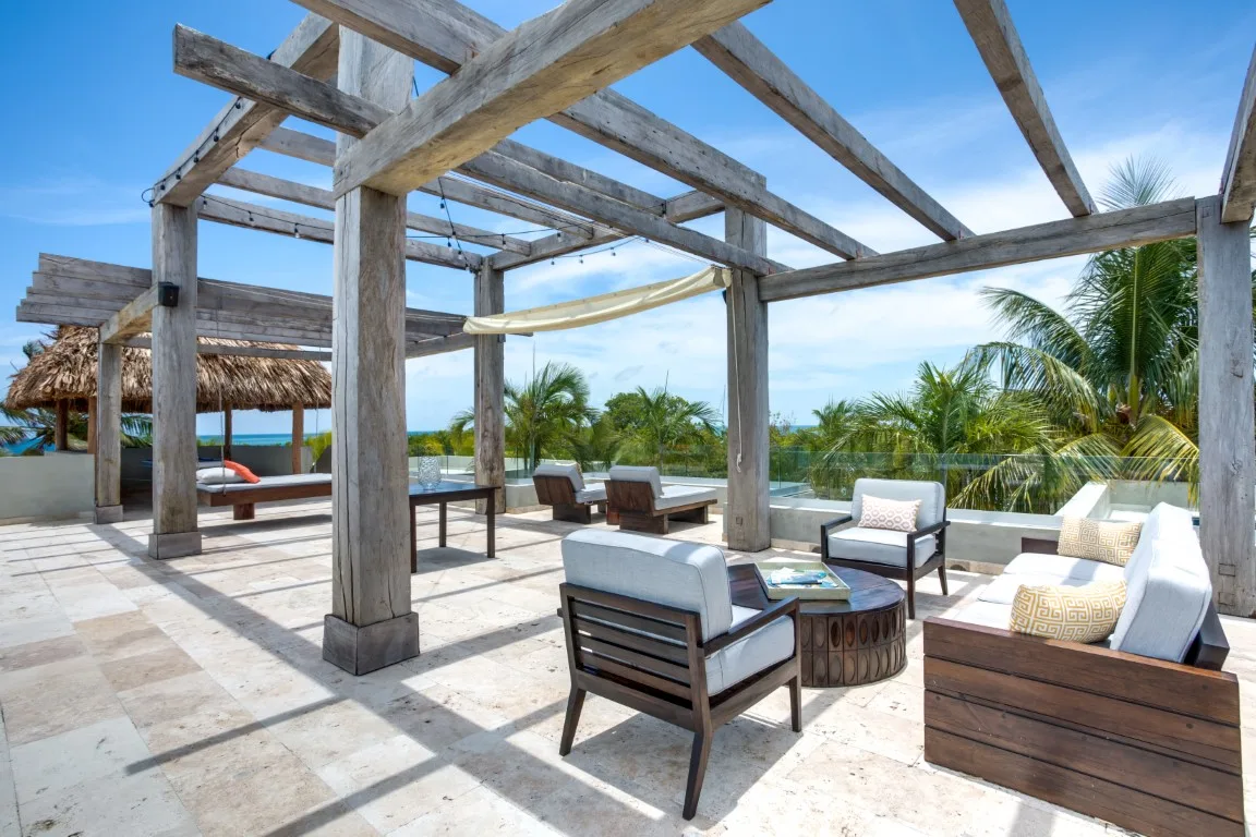 An outdoor patio with seating beneath a wooden pergola, overlooking palm trees.