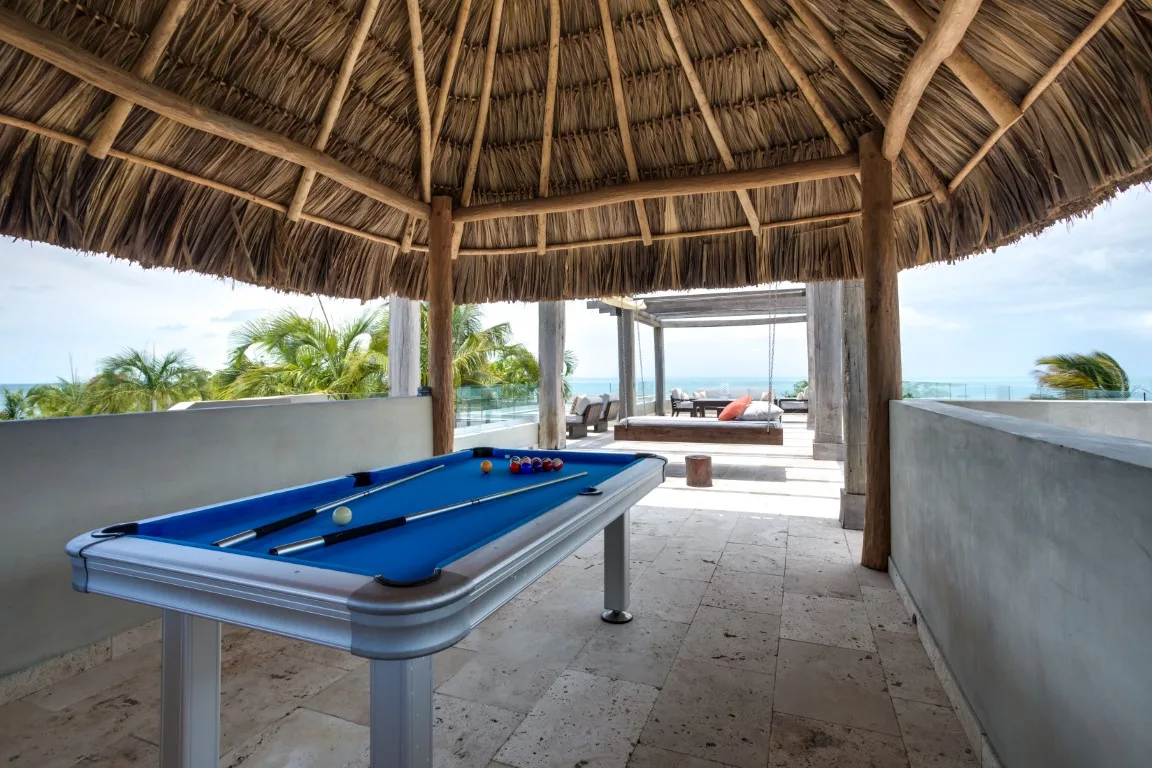 A pool table sits beneath a thatched-roof pavilion overlooking the water.