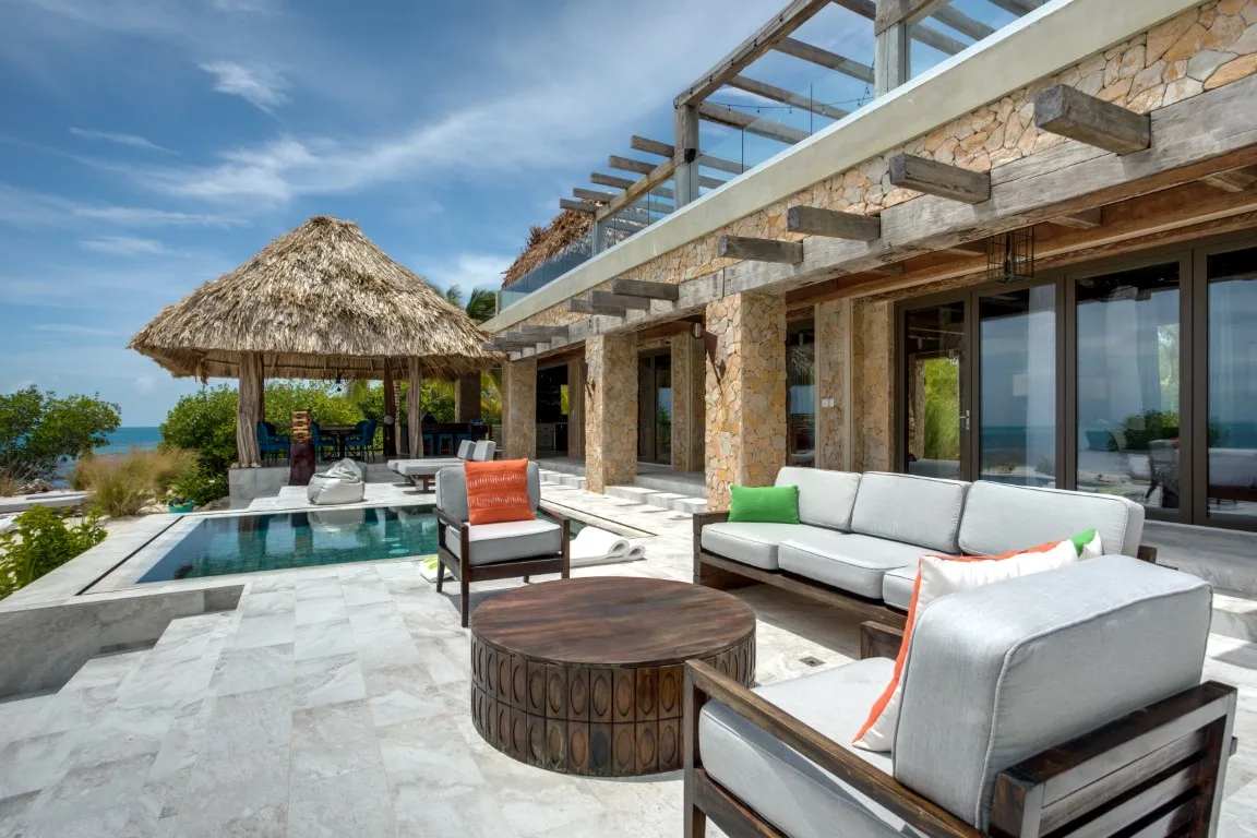 A sunlit outdoor seating area beside a pool, with a thatched-roof pavilion.