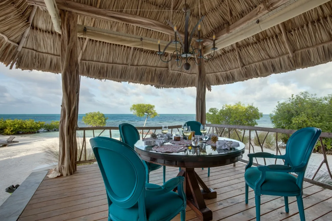 A thatched outdoor dining area overlooks the sea, with a table and turquoise seating.