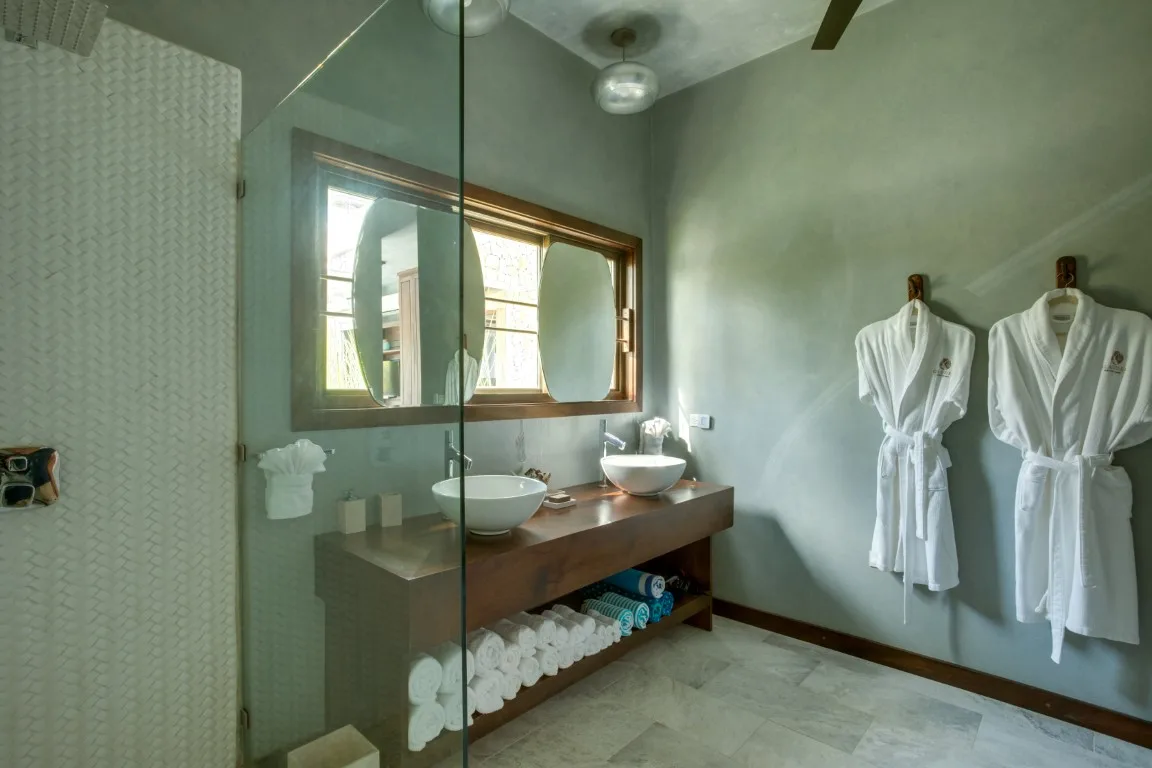 A bathroom vanity and mirror sit beside a glass shower and two hanging robes.