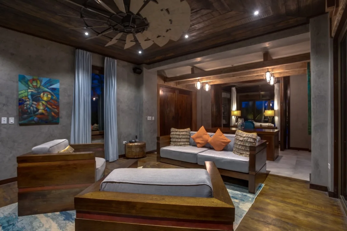 A dimly lit living room has low seating, wooden furnishings, and colorful wall art.