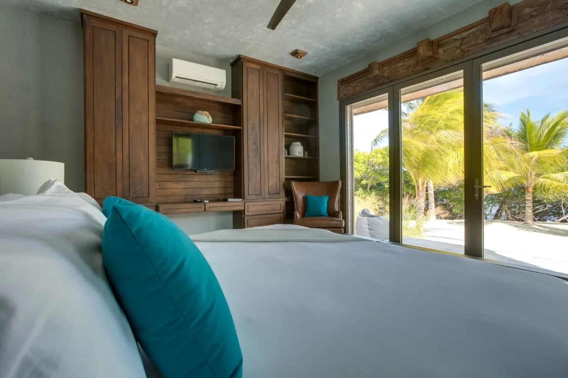 A bedroom with a bed, wooden built-in shelving, and glass doors opening to a palm-lined view.