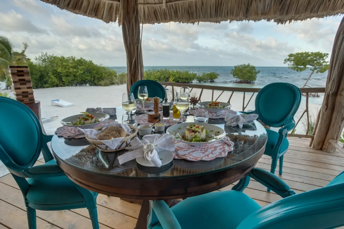 An outdoor dining table is set beneath a thatched roof overlooking the sea.