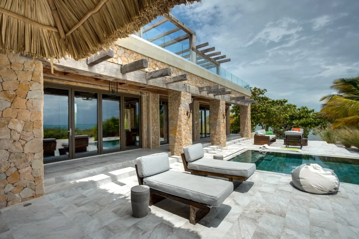 A stone house opens onto a sunlit patio with cushioned seating and a pool.