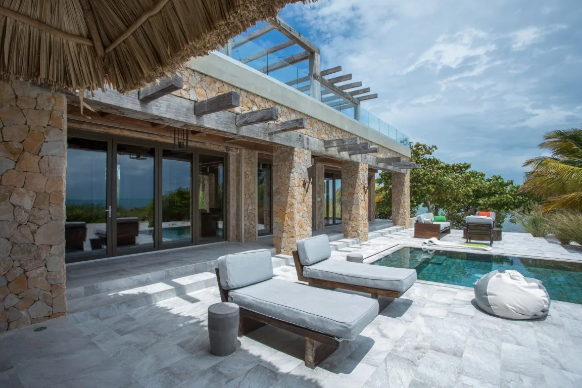 A stone house opens onto a sunlit terrace with lounge seating beside a pool.