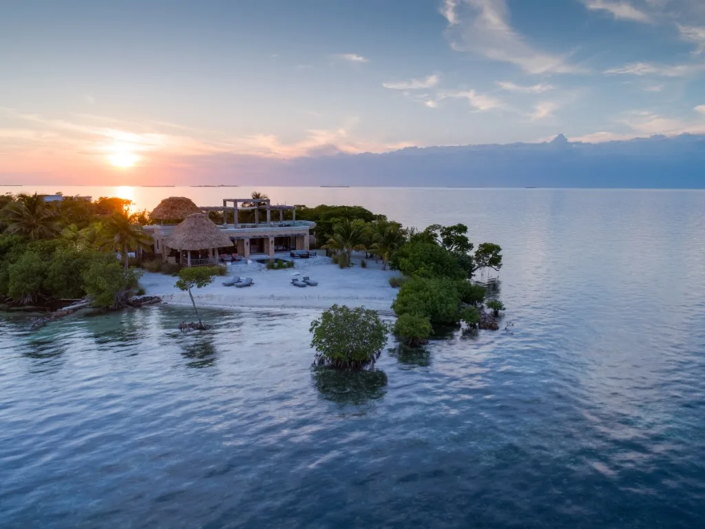 A small tropical island with beachfront buildings sits in calm water at sunset.