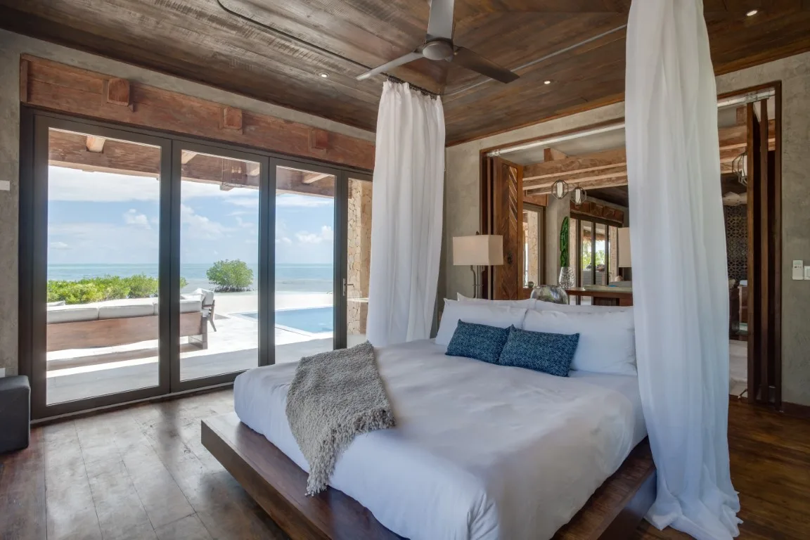 A bedroom with a white bed and curtains beside glass doors overlooking the ocean.