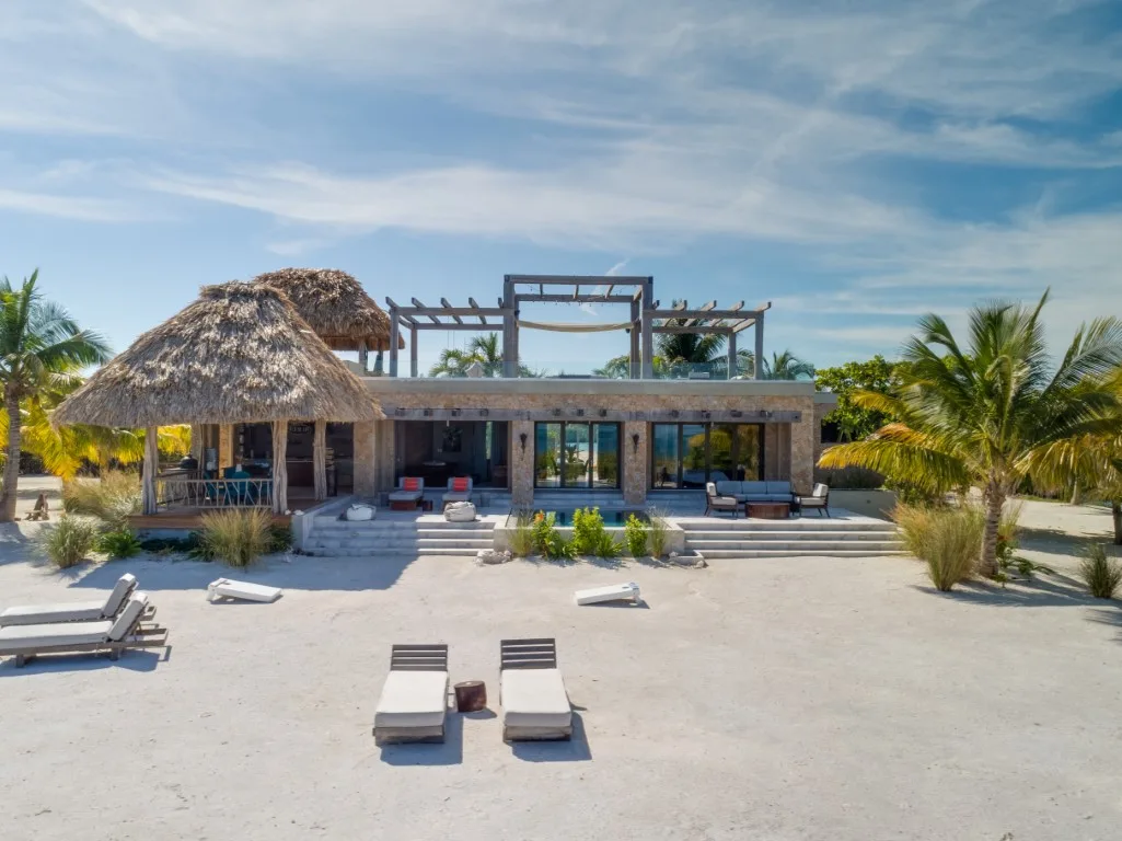 A beachfront-style villa faces a sandy outdoor area with lounge seating and palm trees.